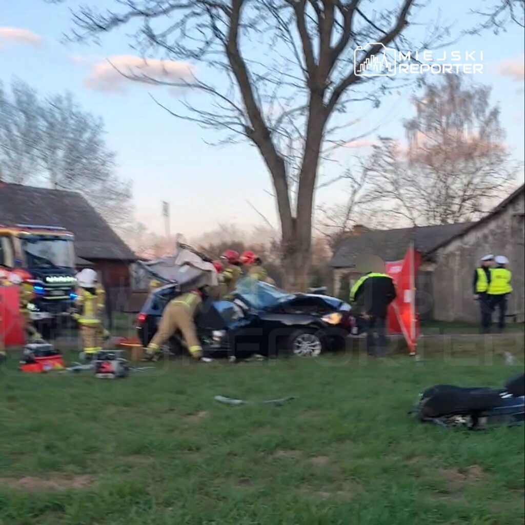 Grupa strażaków pracuje nad wydobyciem poszkodowanego z rozbitego samochodu w wiejskim otoczeniu.