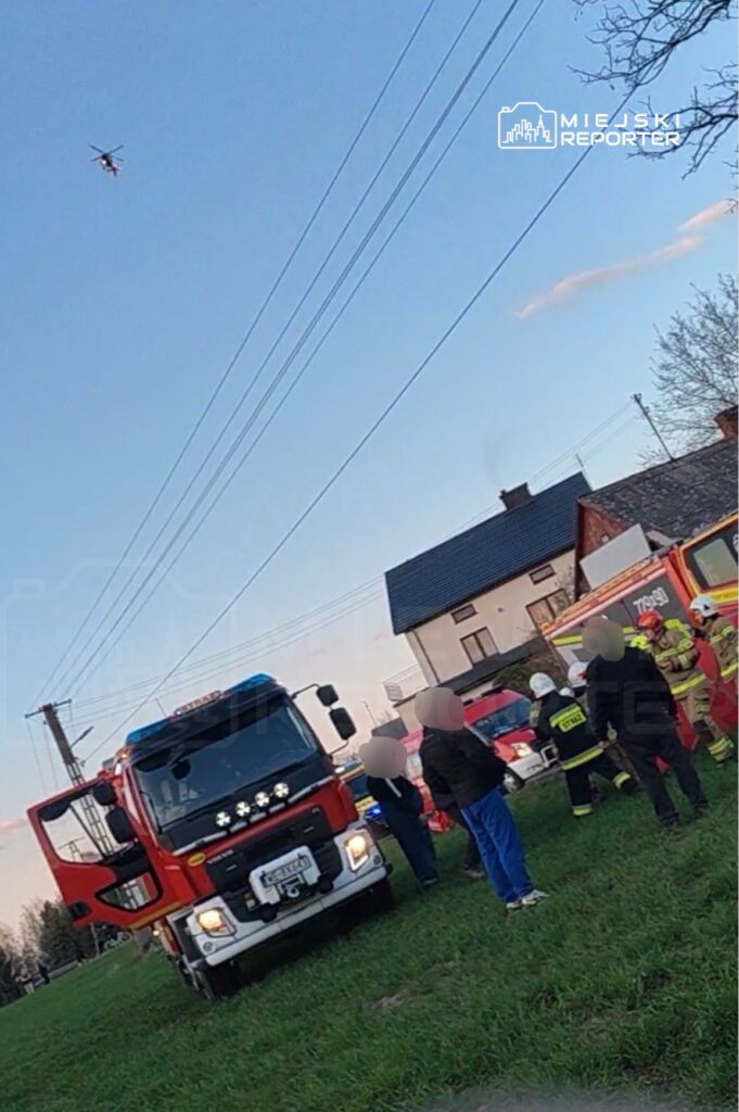 Grupa strażaków w mundurach omawia sytuację obok wozu strażackiego na trawiastym terenie, nad którymi krąży helikopter.
