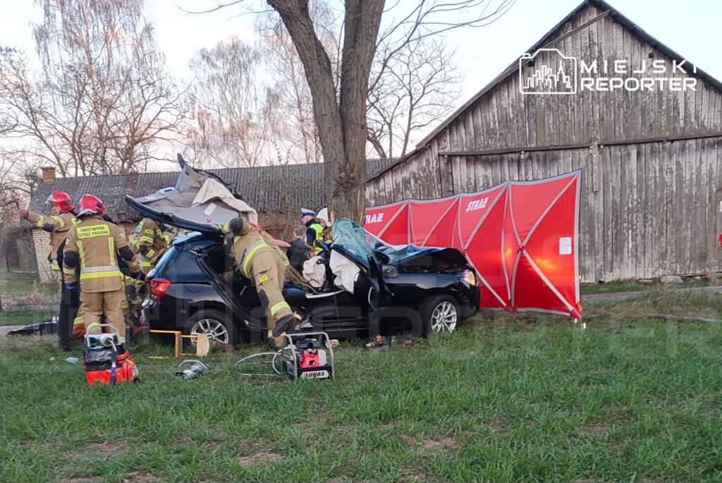 Grupa strażaków w mundurach pracuje przy rozbitej osobówce, w tle widoczna drewniana stodoła.