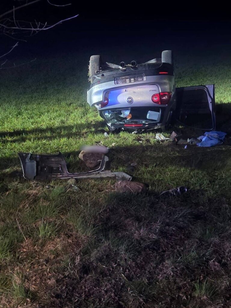 Samochód leżący na dachu na trawiastej powierzchni, obok porozrzucane elementy wnętrza pojazdu.