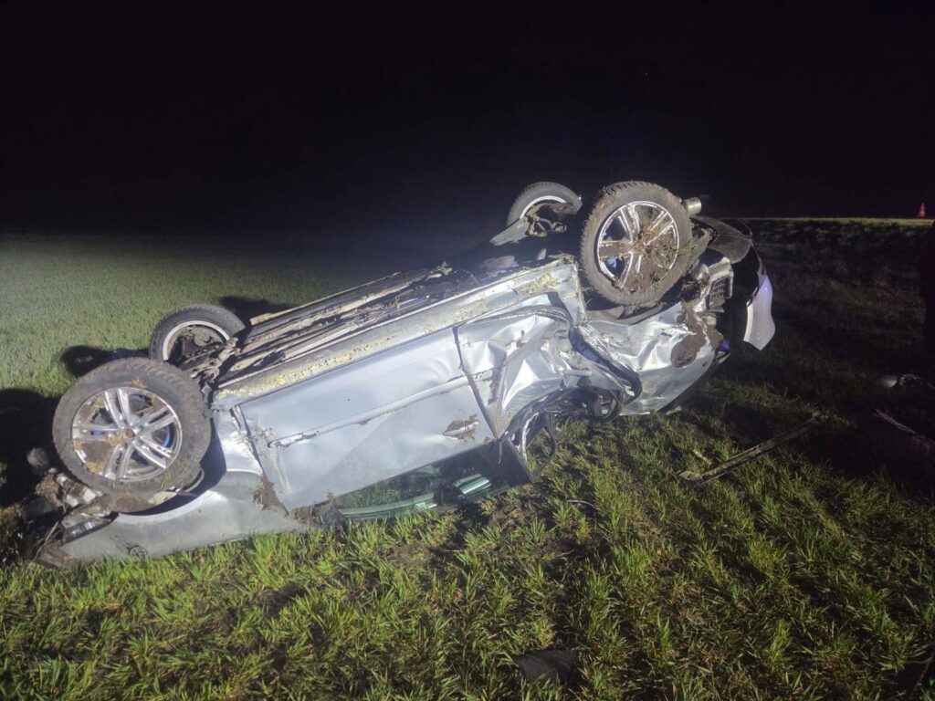 Przewrócony samochód osobowy leży na boku na trawie, w nocy, z widocznymi uszkodzeniami karoserii.