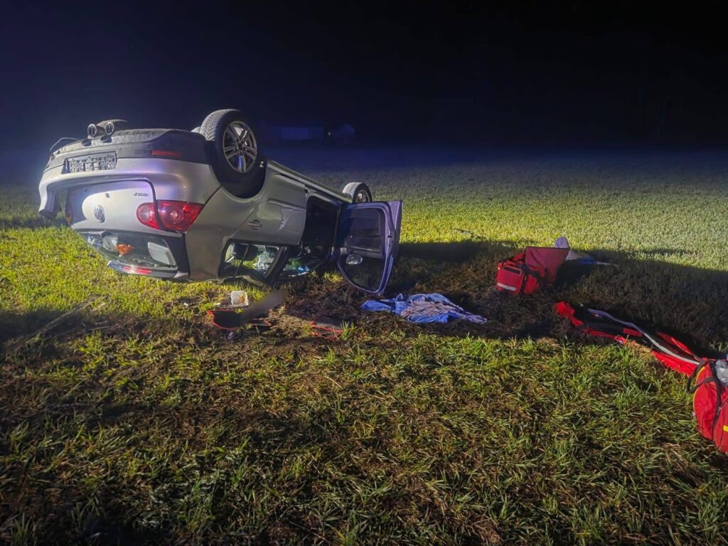 Samochód osobowy leży na dachu na trawie, obok widoczne są porozrzucane przedmioty i torby ratunkowe.