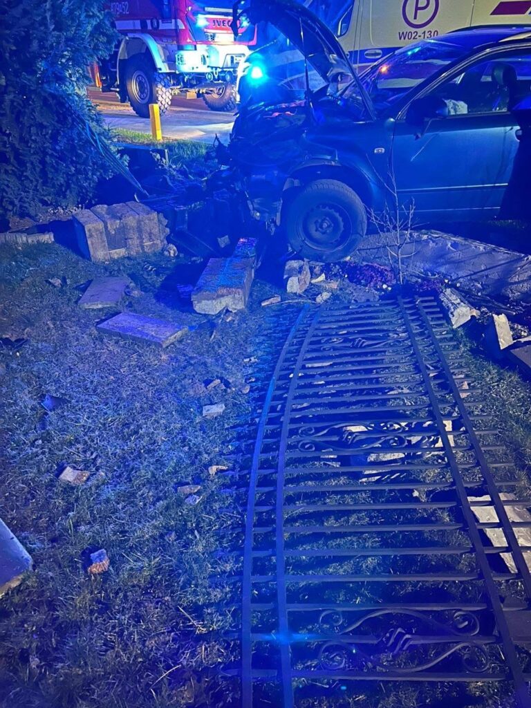 W nocy, niebieskie światła pojazdów ratunkowych oświetlają uszkodzone auto, które wjechało w ogrodzenie, leżące na trawie.