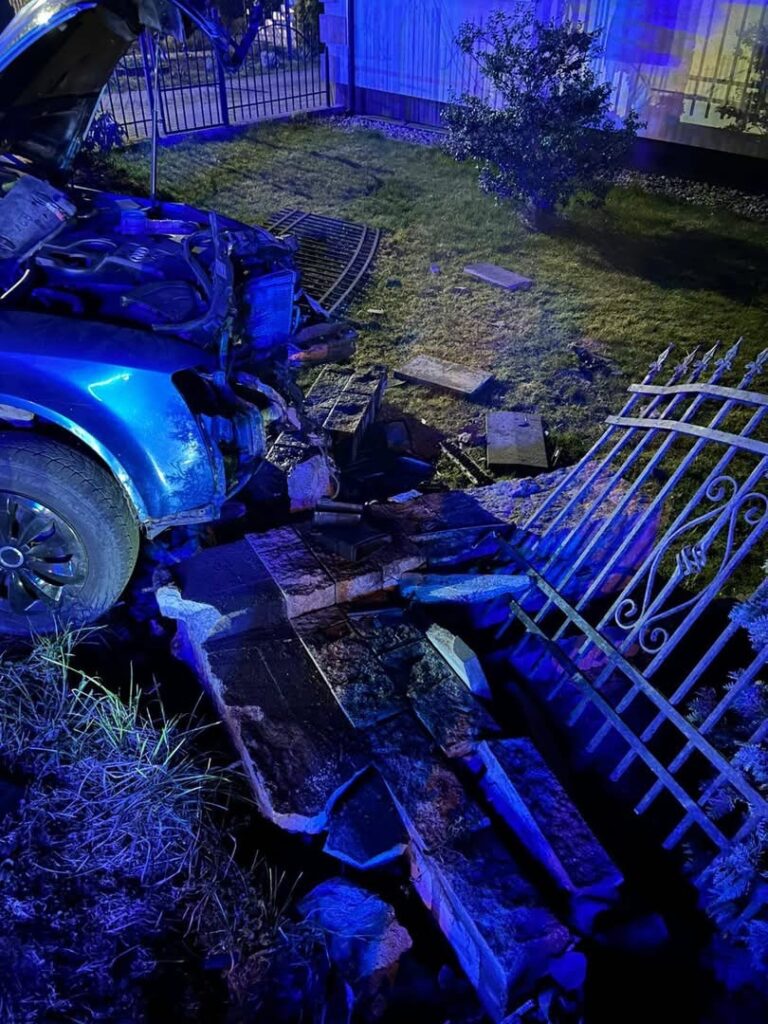 Uszkodzony niebieski samochód stoi na trawniku, zniszczył część ogrodzenia i nawierzchnię przed domem.