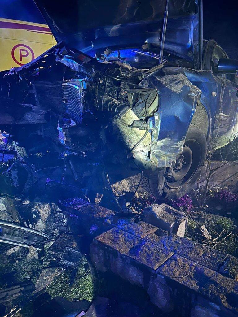 Zniszczony przód samochodu osobowego leży na poboczu drogi, otoczony przez fragmenty muru i trawę, w tle niebieskie światła.