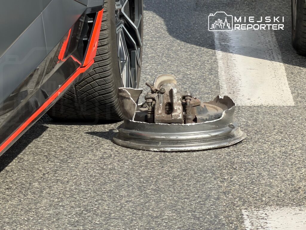 Samochód na parkingu z uszkodzoną felgą leżącą obok, obok widoczna droga z białymi pasami.