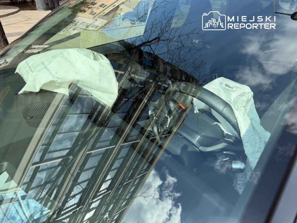 Wnętrze samochodu z widocznym poduszką powietrzną na kierownicy, kawałek materiału leży na desce rozdzielczej.