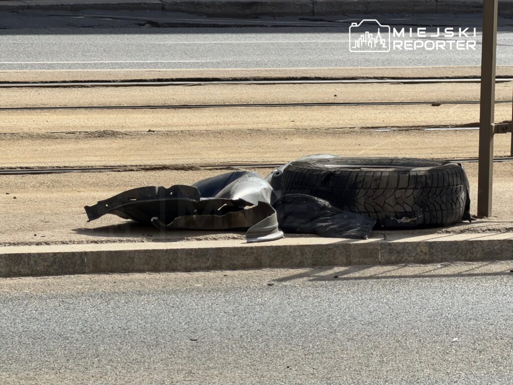 Na chodniku leży uszkodzona opona oraz fragmenty plastikowych elementów samochodowych.