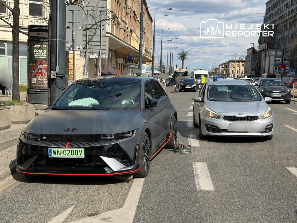 Czarny samochód osobowy z uszkodzoną oponą stoi na jezdni, obok srebrny pojazd policyjny z włączonymi sygnałami świetlnymi.