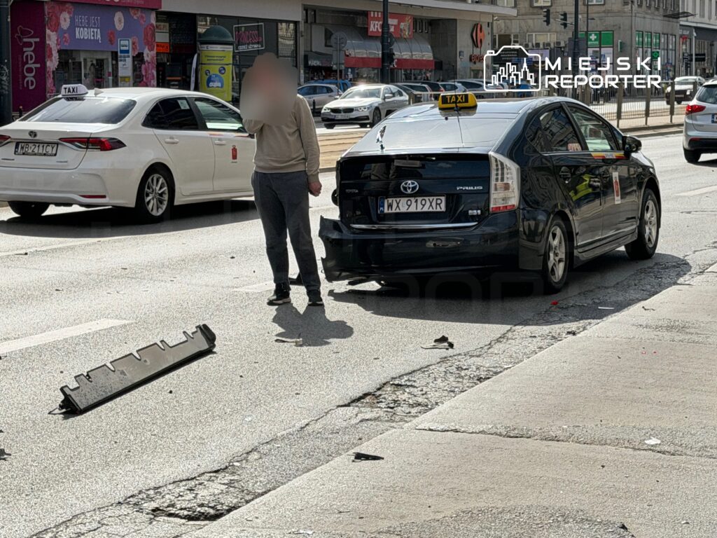 Mężczyzna w szarym swetrze stoi na drodze obok uszkodzonego czarnego taksówki, a w tle widoczna biała taksówka.