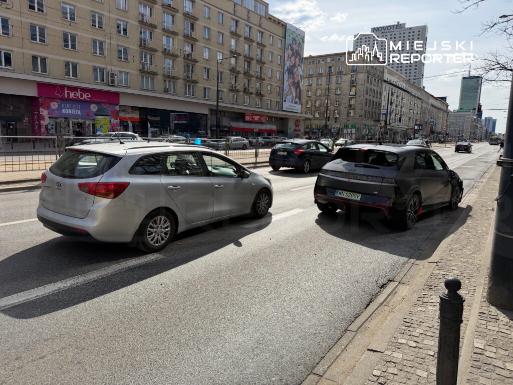 Dwa samochody zaparkowane na ulicy, obok nowoczesnych budynków w tle, z widocznymi reklamami na fasadach.