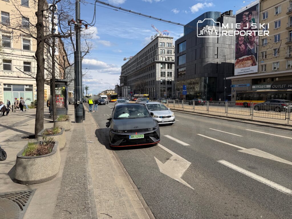 Ruch uliczny w mieście z samochodami na jezdni, przechodniami na chodniku oraz nowoczesnym biurowcem w tle.