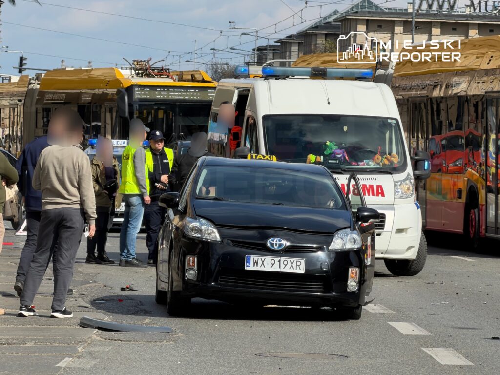 Grupa osób w żółtych kamizelkach rozmawia z policjantem obok zderzonego taksówki i karetki na ulicy.