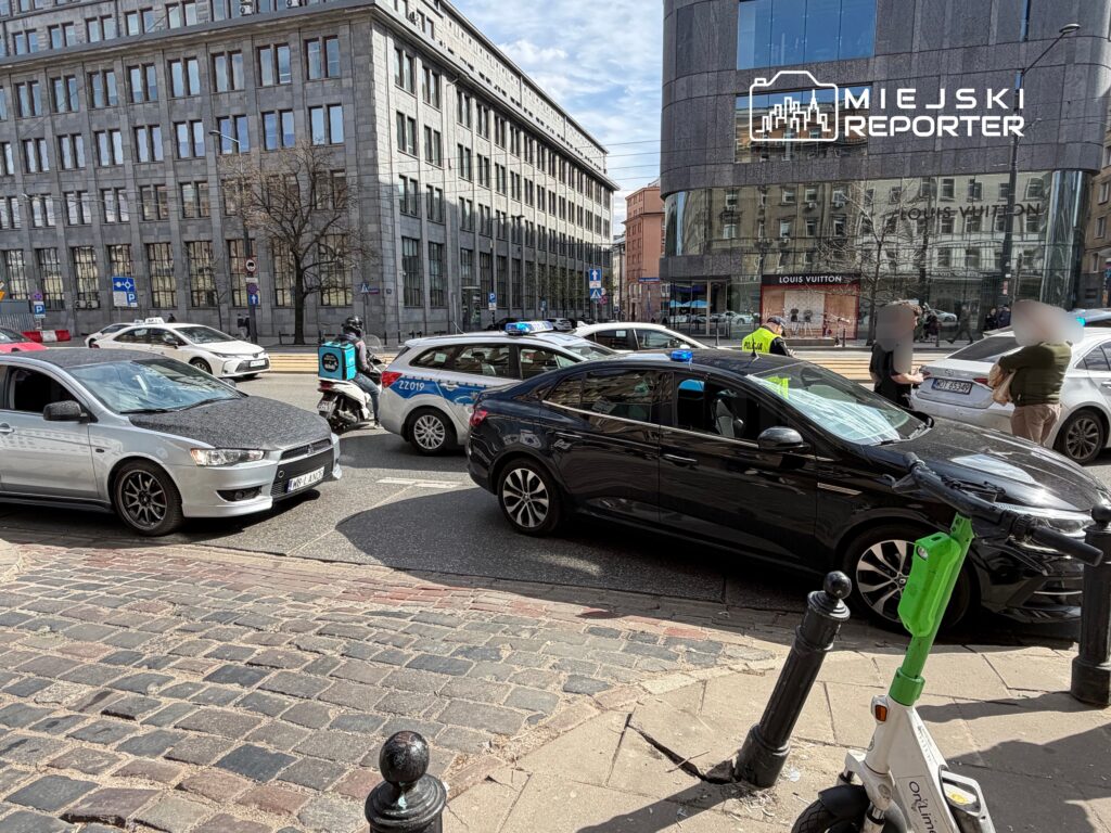 Kierowca w czarnym samochodzie stoi z policjantem obok, podczas gdy inne pojazdy czekają w ruchu ulicznym.