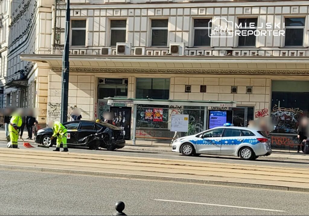 Na ulicy, pracownicy w odblaskowych kamizelkach sprzątają miejsce wypadku, obok stoi uszkodzony samochód oraz radiowóz policji.