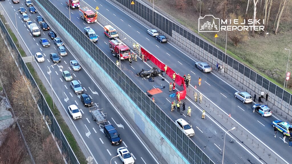 Na jezdni leżą wrak przewróconego samochodu, a obok pracują strażacy i policjanci, zabezpieczając miejsce wypadku.