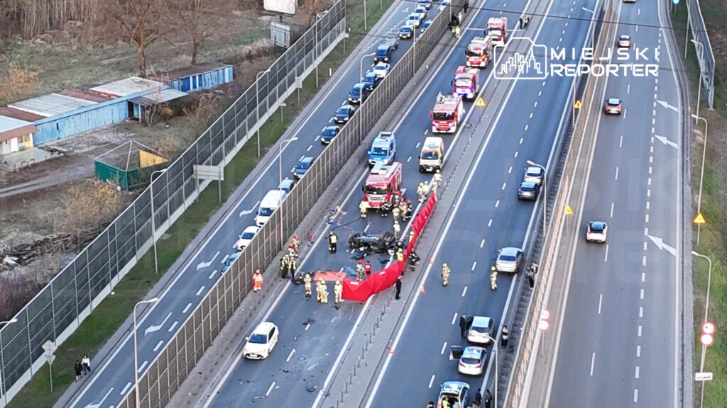 Na jezdni leżą wrak samochodu oraz rozstawione bariery, wokół pracują strażacy i policjanci.
