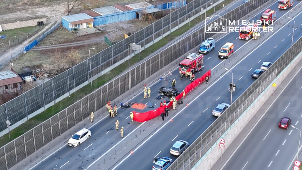 Grupa strażaków i ratowników medycznych pracuje na miejscu wypadku, zabezpieczając uszkodzony pojazd na drodze.