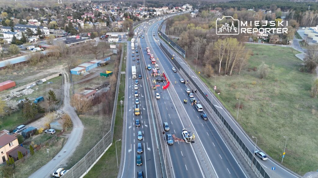 Na drodze widać kolizję samochodów oraz interwencję służb ratunkowych. Ruch jest zablokowany w obu kierunkach.
