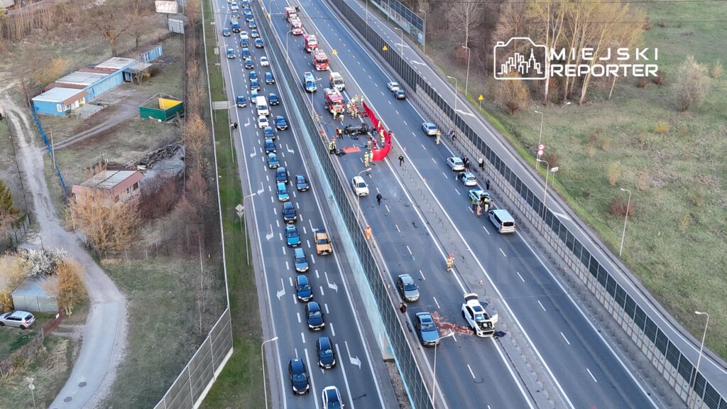 Na jezdni dużego ruchu drogowego, zespoły ratunkowe pracują przy wypadku samochodowym, zabezpieczając miejsce zdarzenia.