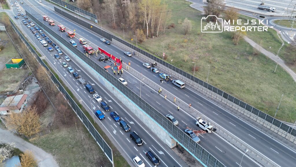 Ruch drogowy zatrzymany na jezdni, gdzie służby ratunkowe pracują przy wypadku samochodowym. W tle widoczne pojazdy.