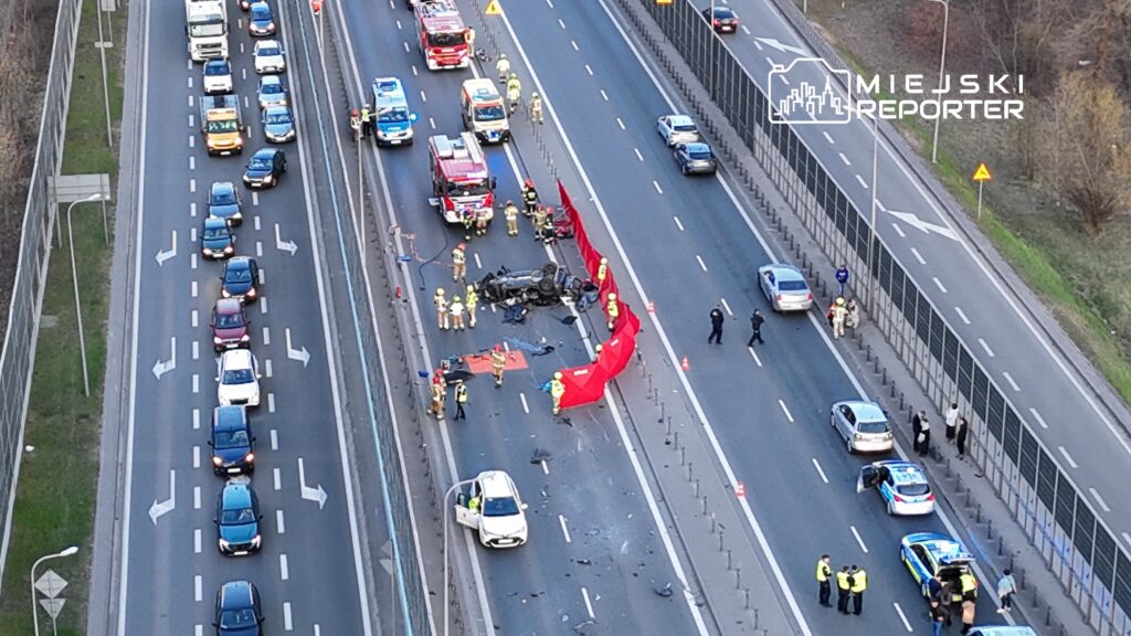 Wielu ratowników medycznych i strażaków pracuje przy przewróconym samochodzie na autostradzie, zabezpieczając miejsce zdarzenia.