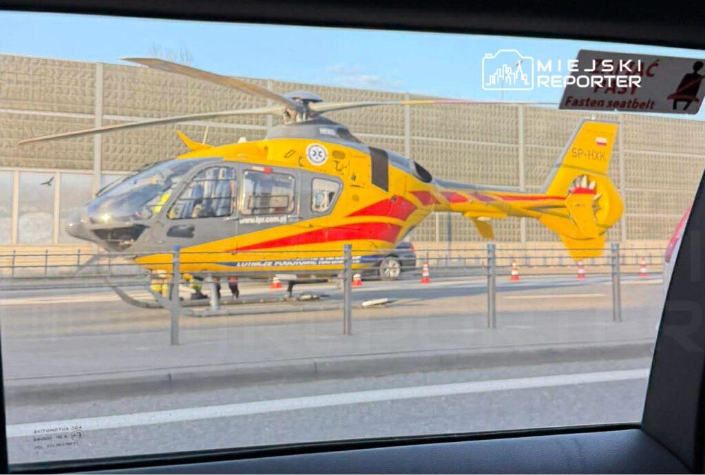 Śmigłowiec ratunkowy w kolorach żółtym i czerwonym stoi na pasie drogowym obok ogrodzenia.