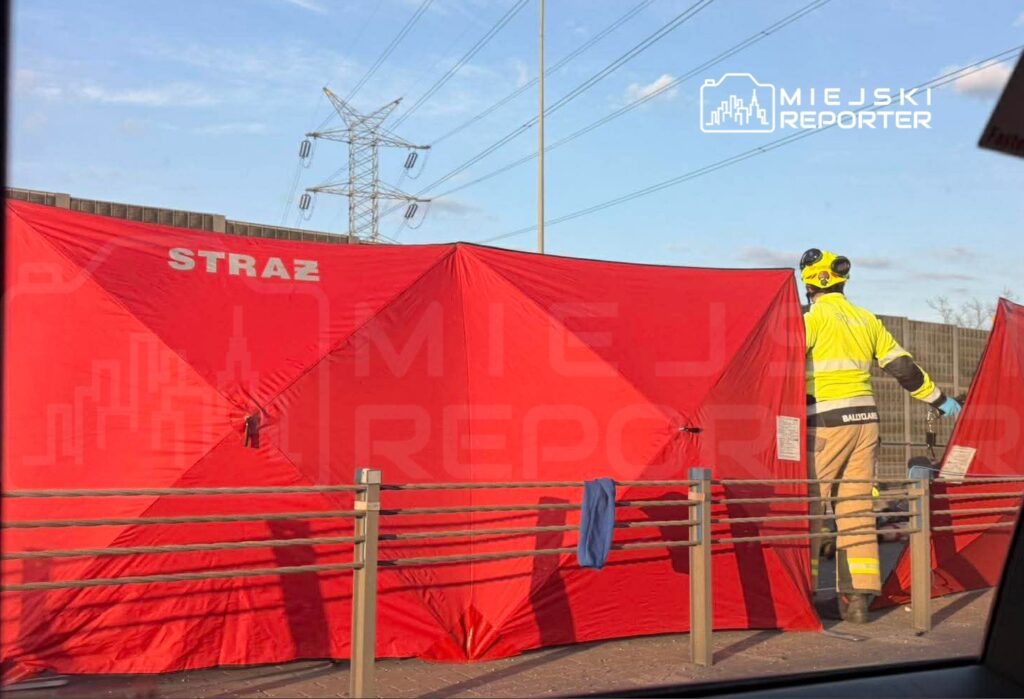 Strażak w żółtej odzieży ochronnej sprawdza czerwony namiot z napisem "STRAŻ" w miejskim otoczeniu.