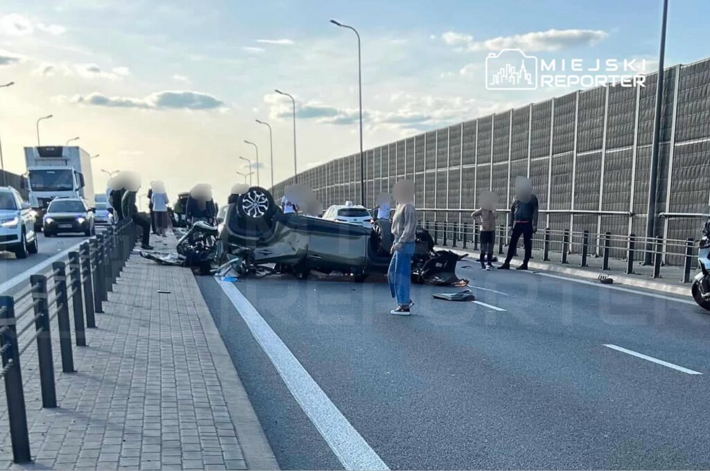 Na autostradzie leży przewrócone auto, a wokół zbierają się ludzie, obserwując sytuację.