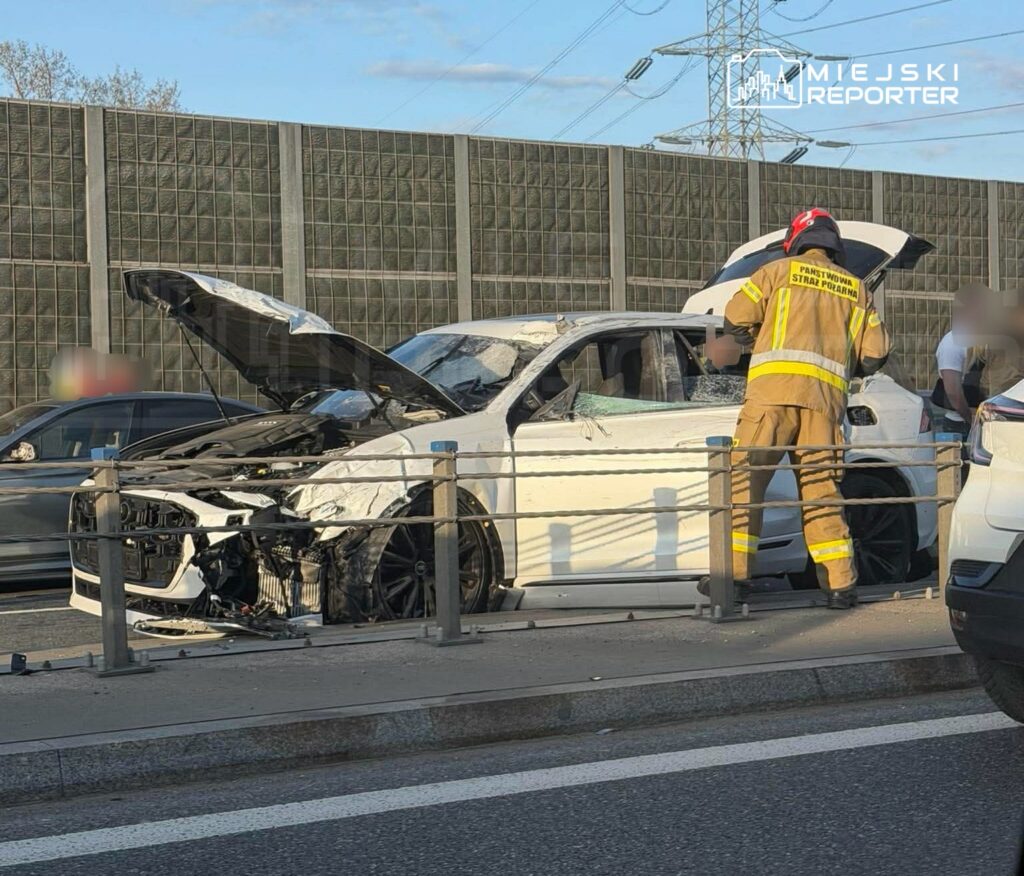 Strażak w mundurze przeszukuje uszkodzony samochód osobowy na drodze, obok stoją inne pojazdy.