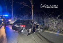 Czarny samochód osobowy z uszkodzeniami stoi na drodze, obok grupy strażaków i pojazdu ratunkowego w nocy.