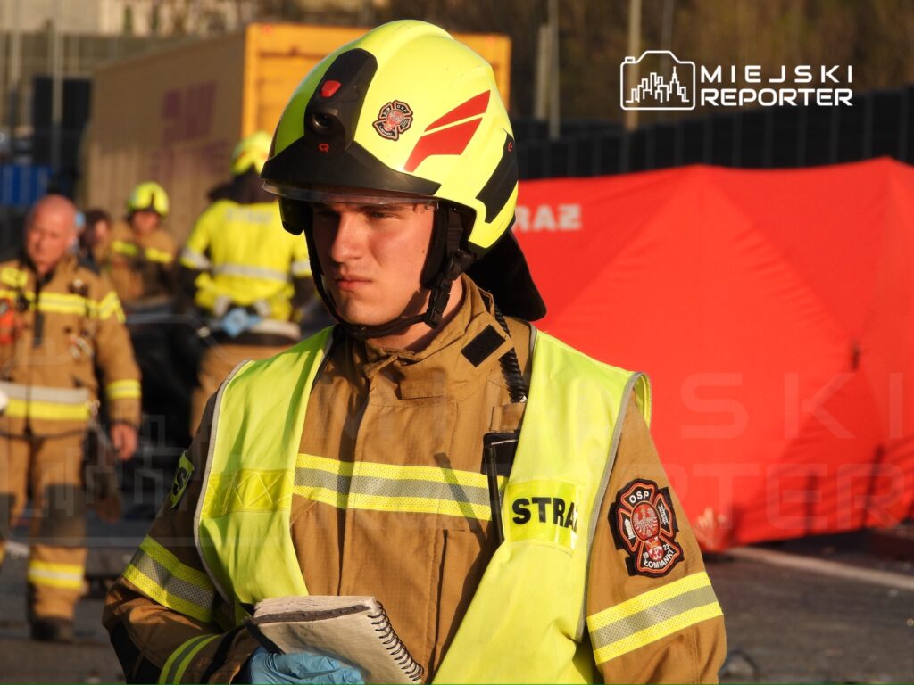 Młody strażak w hełmie i mundurze z notatnikiem w ręku, obserwujący akcję ratunkową w tle.