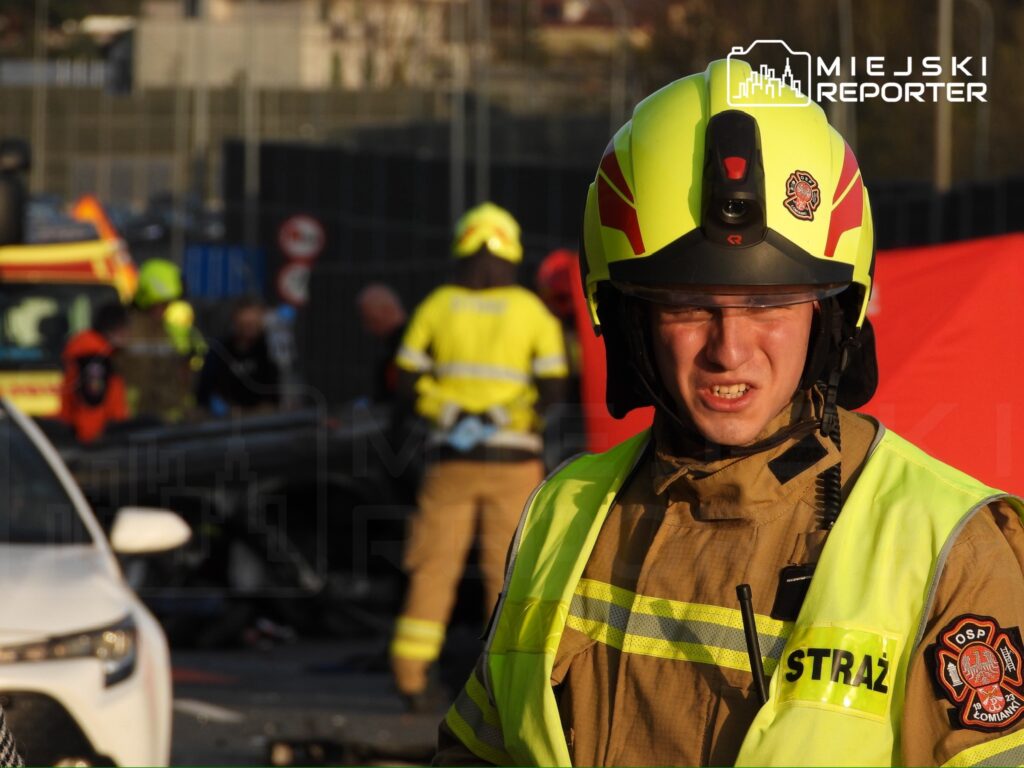 Strażak w hełmie i mundurze, z wyrazem skupienia, stoi na miejscu wypadku, w tle zespół ratunkowy.