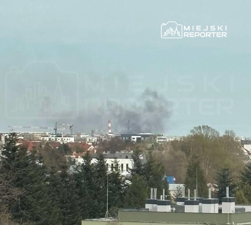 Czarny dym unoszący się nad miejskim krajobrazem w tle, z widocznymi budynkami i drzewami w pierwszym planie.