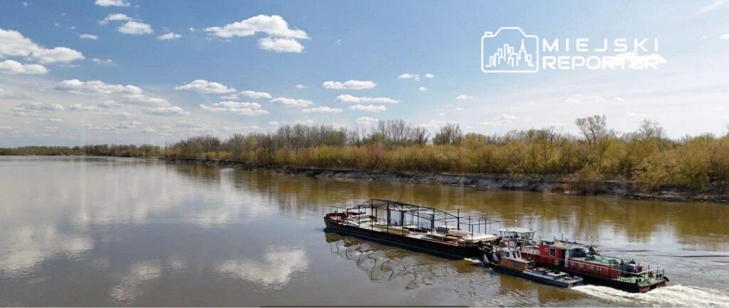Barka transportowa przepływająca po rzece w otoczeniu zielonych brzegów i błękitnego nieba z chmurami.