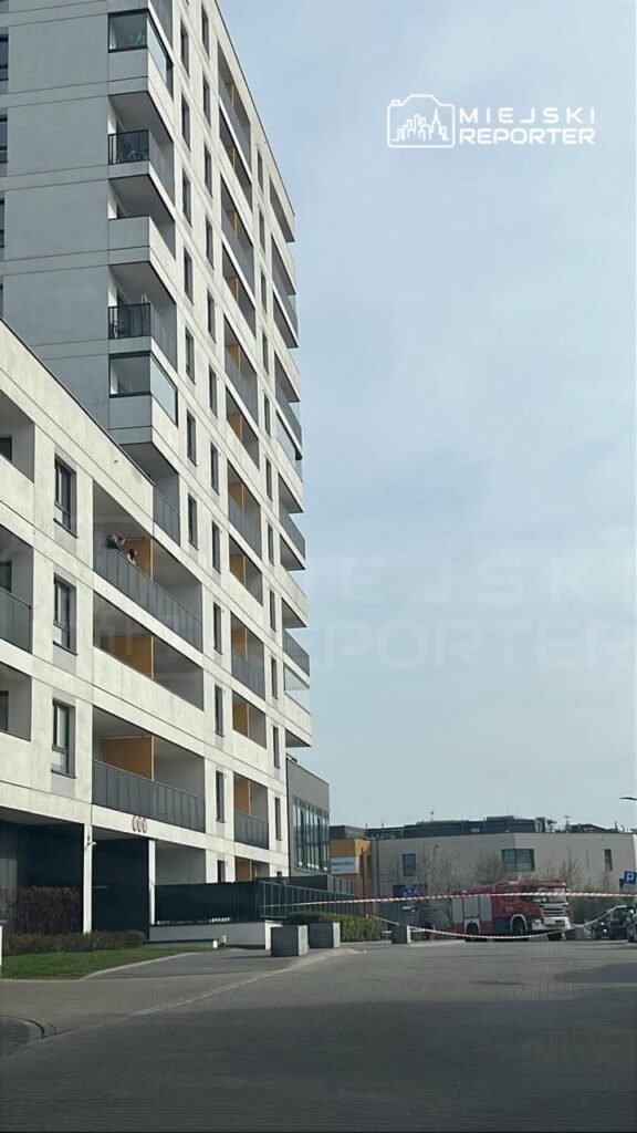 Nowoczesny budynek mieszkalny z balkonami, w tle woz strażacki i taśma zabezpieczająca, na podjeździe.