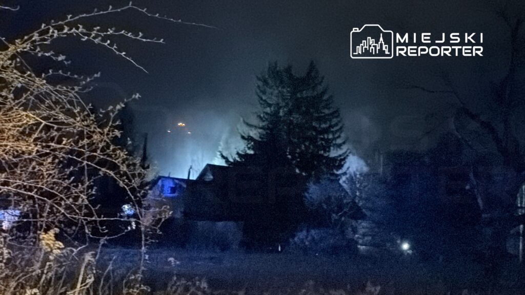 Widok na ciemny krajobraz nocny z oświetlonymi budynkami i drzewami w tle, w tle dym unoszący się w powietrzu.