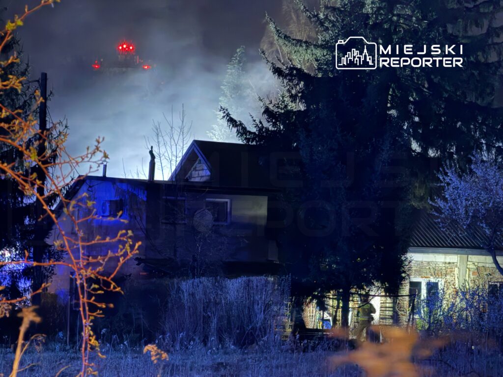 Czerwone światła pojazdu strażackiego oświetlają dymiący dom w otoczeniu drzew.