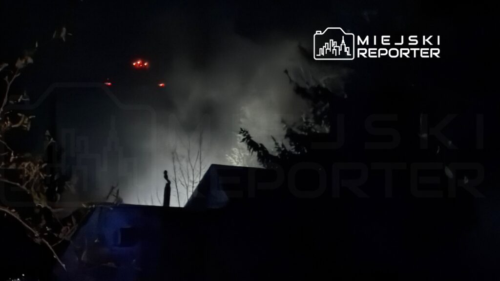 Dym wydobywający się z budynku w nocy, z oświetleniem w tle i zarysem drzew.