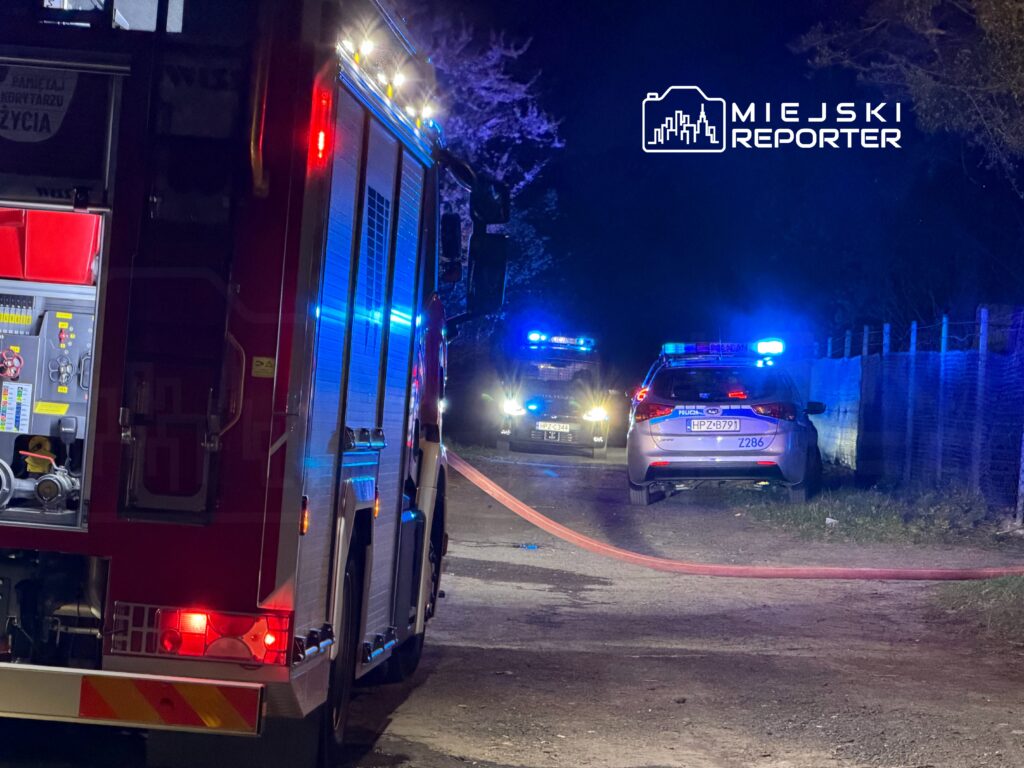W nocy, w ciemnej okolicy, widać wóz strażacki oraz radiowozy policyjne z włączonymi sygnałami świetlnymi.