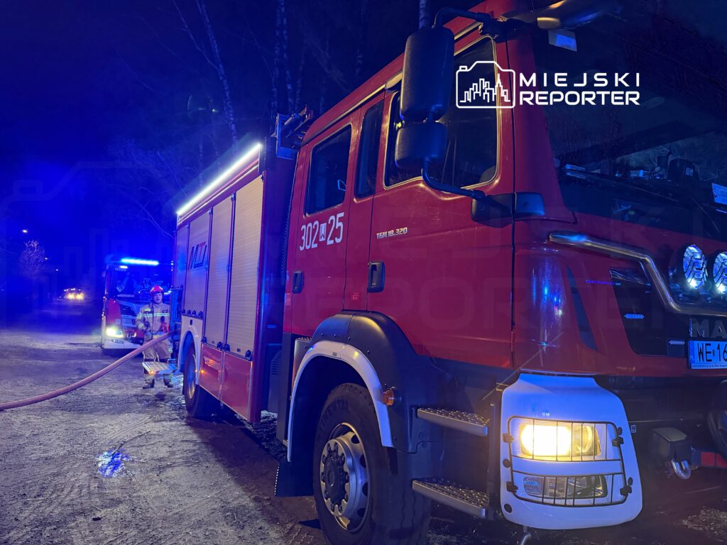 Strażak w hełmie rozciąga wąż gaśniczy obok wozu strażackiego na oświetlonej niebieskim światłem drodze.