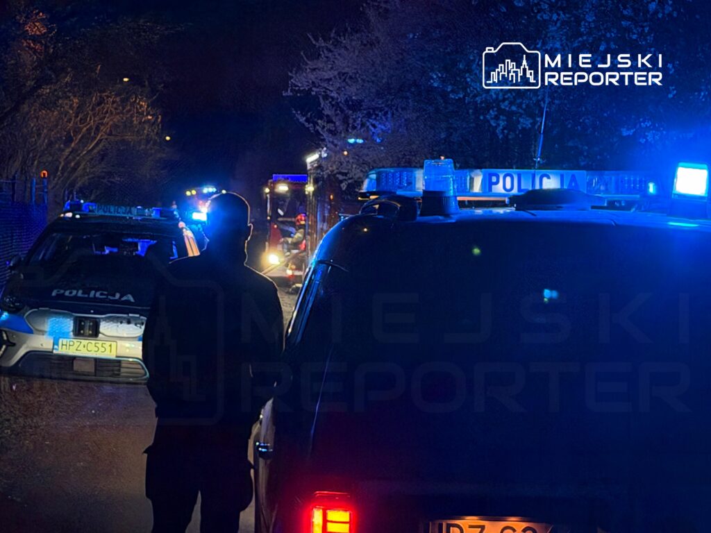 Policjant obserwujący akcję ratunkową w nocy, w otoczeniu pojazdów straży pożarnej i policyjnych z włączonymi sygnałami.