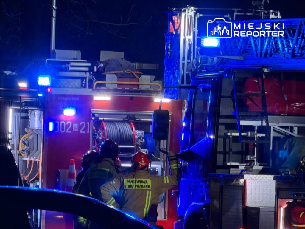 Strażacy w hełmach i uniformach omawiają sytuację przy wozie strażackim z włączonymi sygnalizatorami świetlnymi.