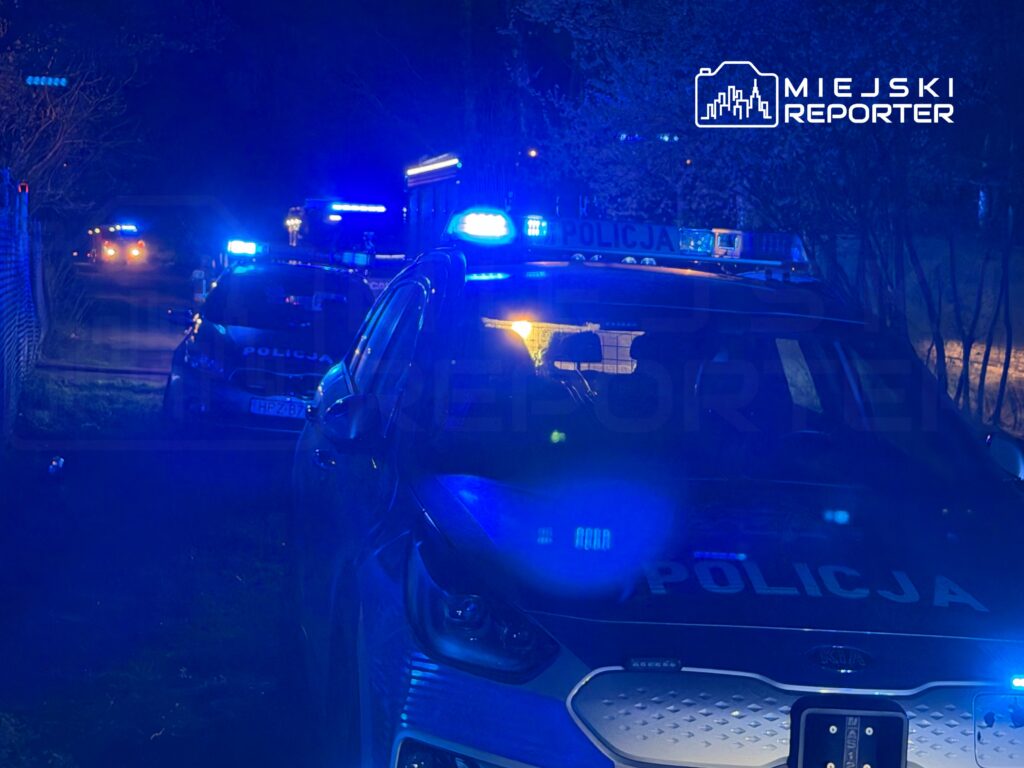 Nocna akcja policji z niebieskimi światłami w tle, pojazdy policyjne zaparkowane w zaciemnionej okolicy.