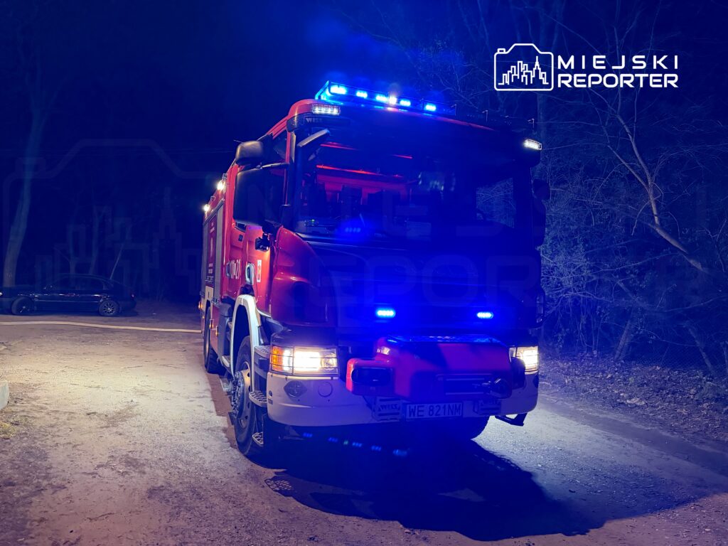 Czerwony wóz strażacki z włączonymi sygnalizatorami świetlnymi stoi na oświetlonej drodze w lesie nocą.