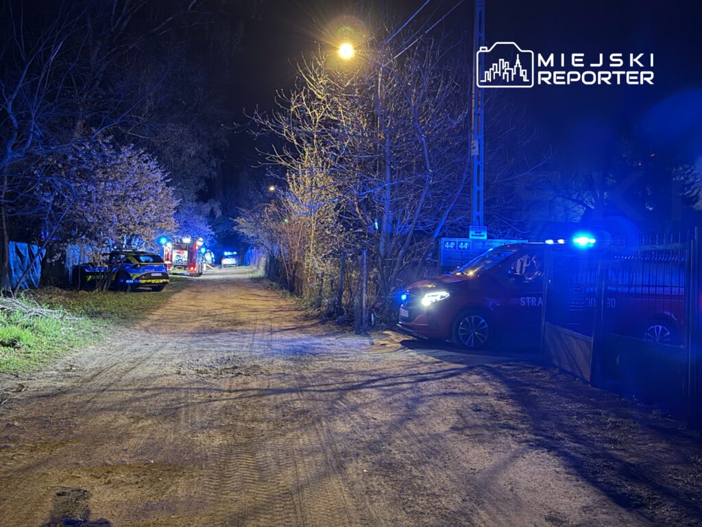 Na wąskiej, oświetlonej drodze nocą, zaparkowane pojazdy służb ratunkowych z włączonymi sygnałami świetlnymi.