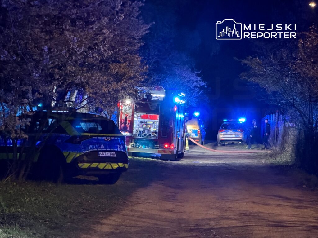 Na wąskiej drodze w nocy, pojazdy policyjne i strażackie z włączonymi sygnałami świetlnymi, w otoczeniu drzew.
