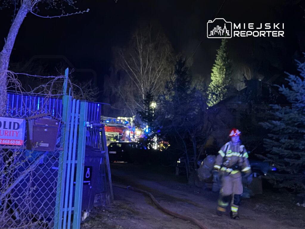 Strażak w mundurze z hełmem przechodzi obok ogrodzenia, w tle widoczne pojazdy strażackie i oświetlenie akcji ratunkowej.