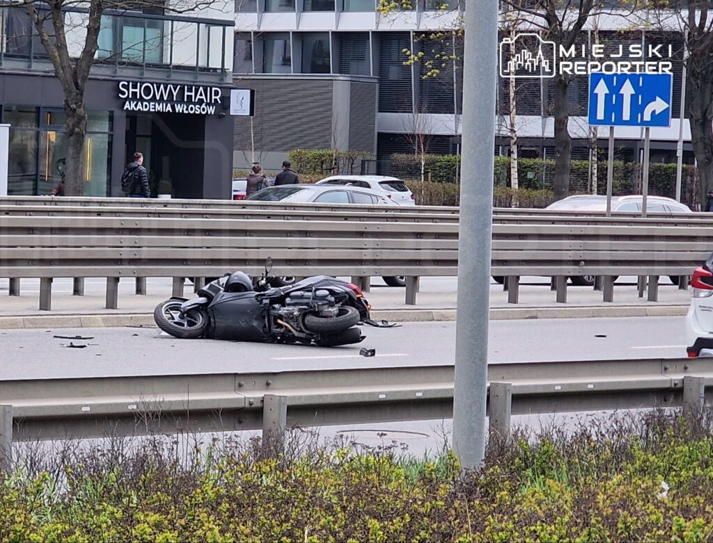 Motocykl leży na jezdni, obok widoczne uszkodzenia, w tle nowoczesny budynek oraz przechodnie.