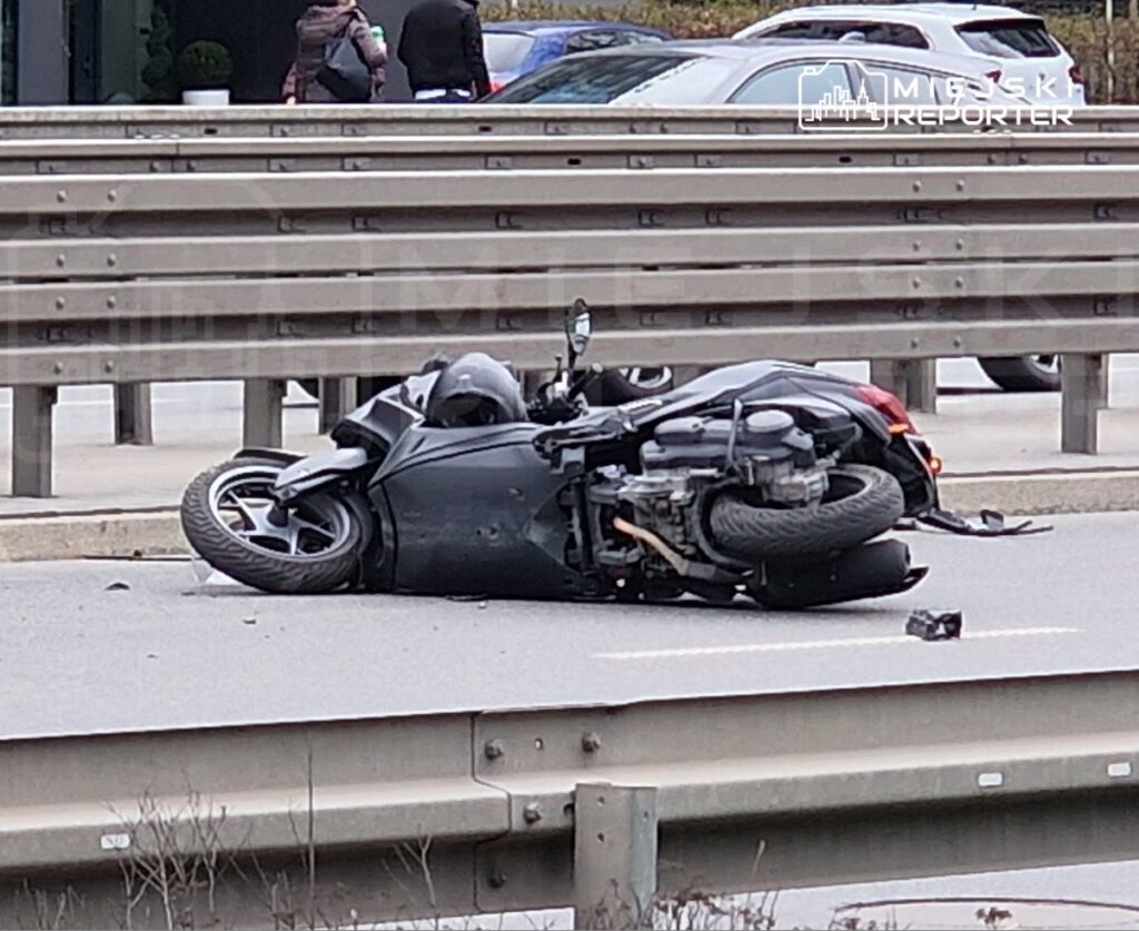 Motocykl leży na boku na jezdni, obok widoczne samochody oraz barierka ochronna. Na drodze dostrzegalne fragmenty.